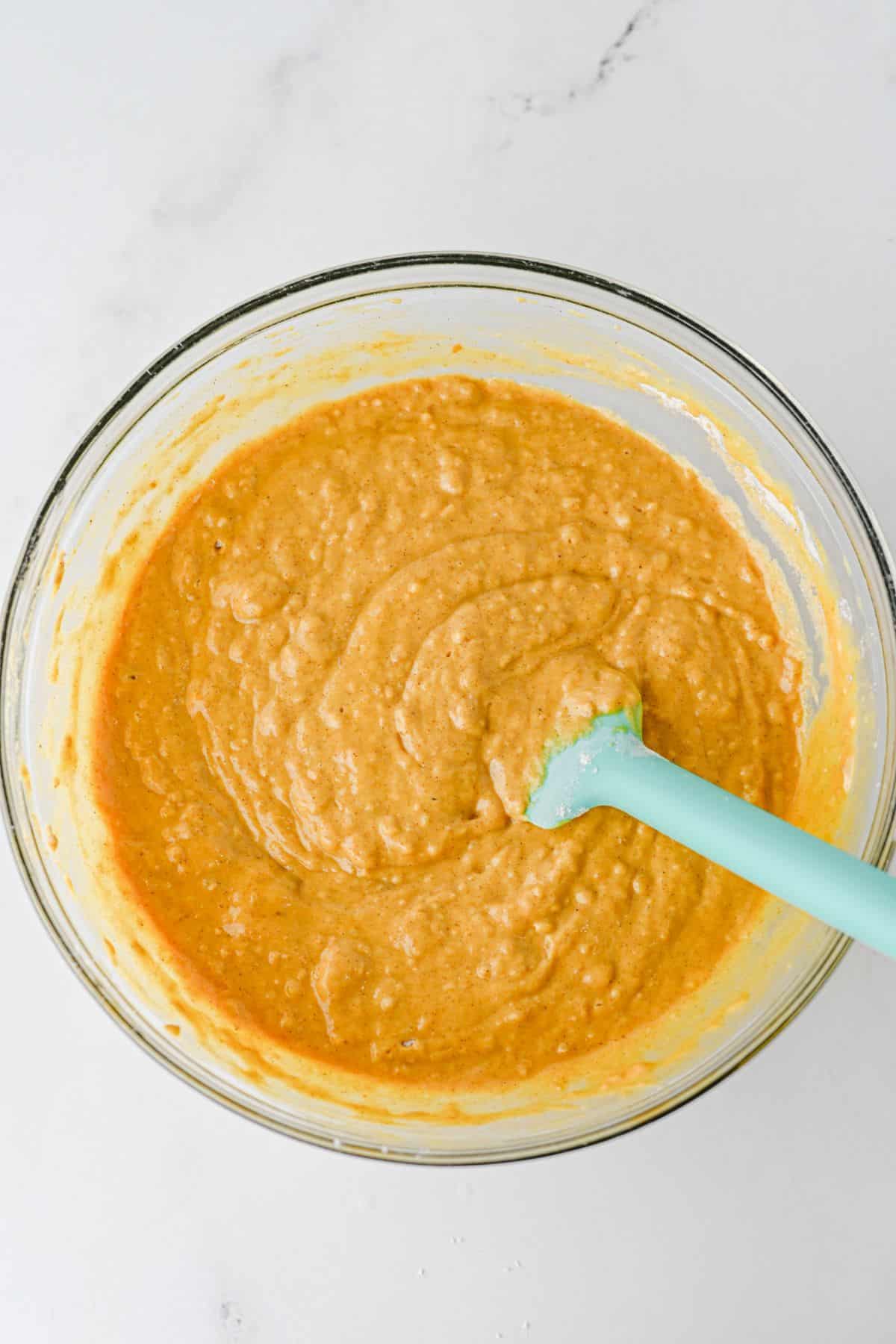 A blue silicone spatula mixes orange cake batter in a clear glass bowl.