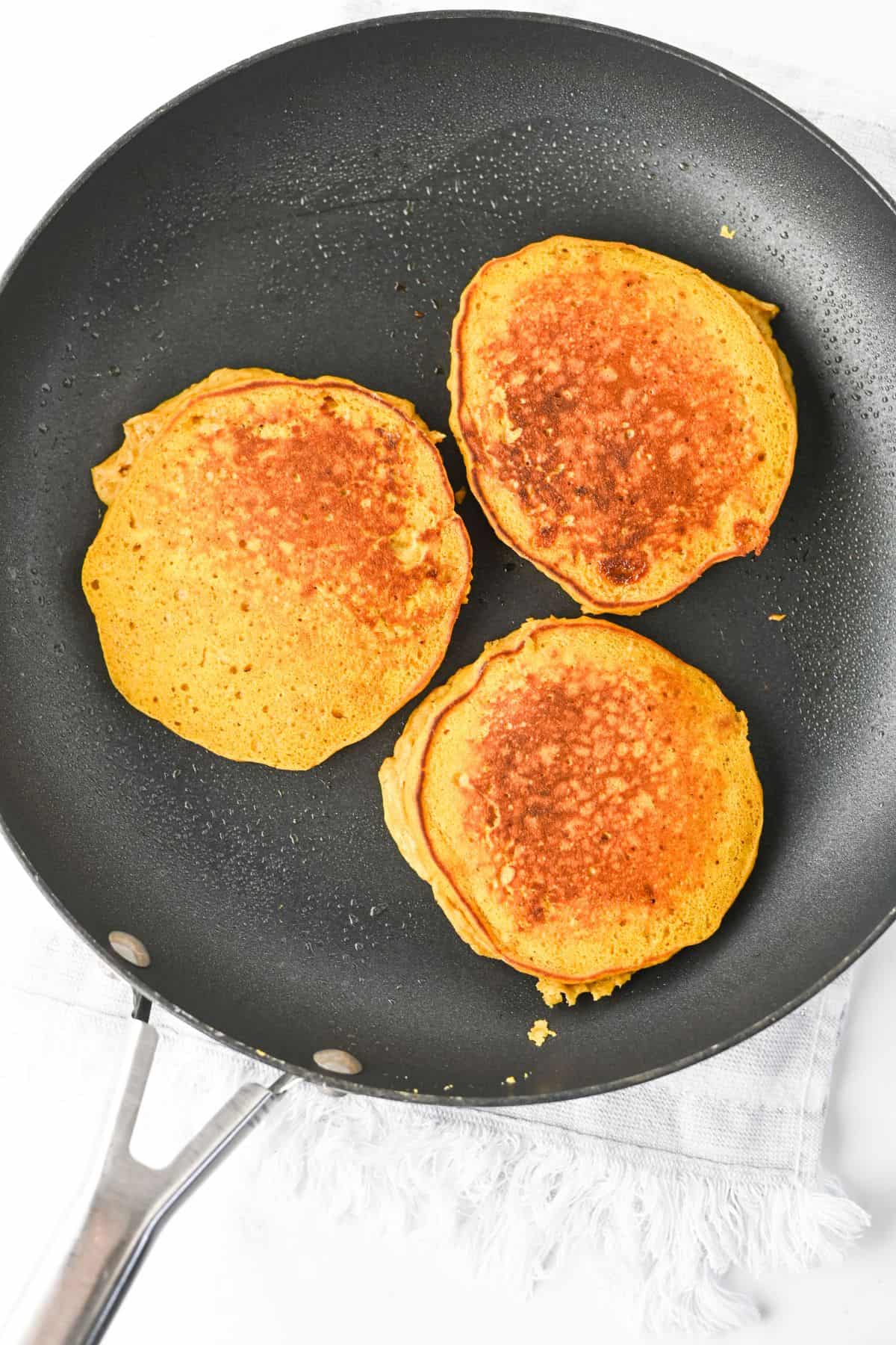 Three golden-brown pancakes cooking in a black frying pan.