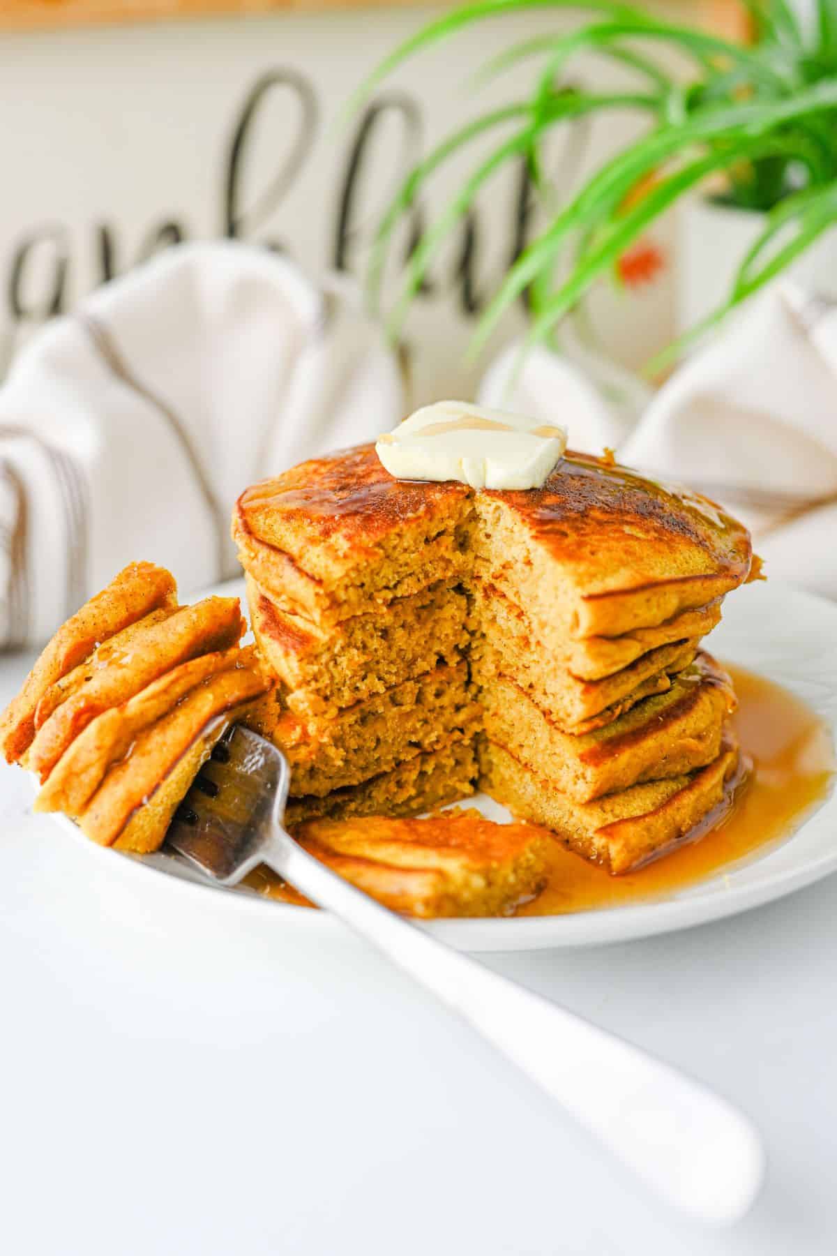 A stack of pumpkin pancakes on a plate topped with a pat of butter and syrup, with a fork holding a piece from the stack.