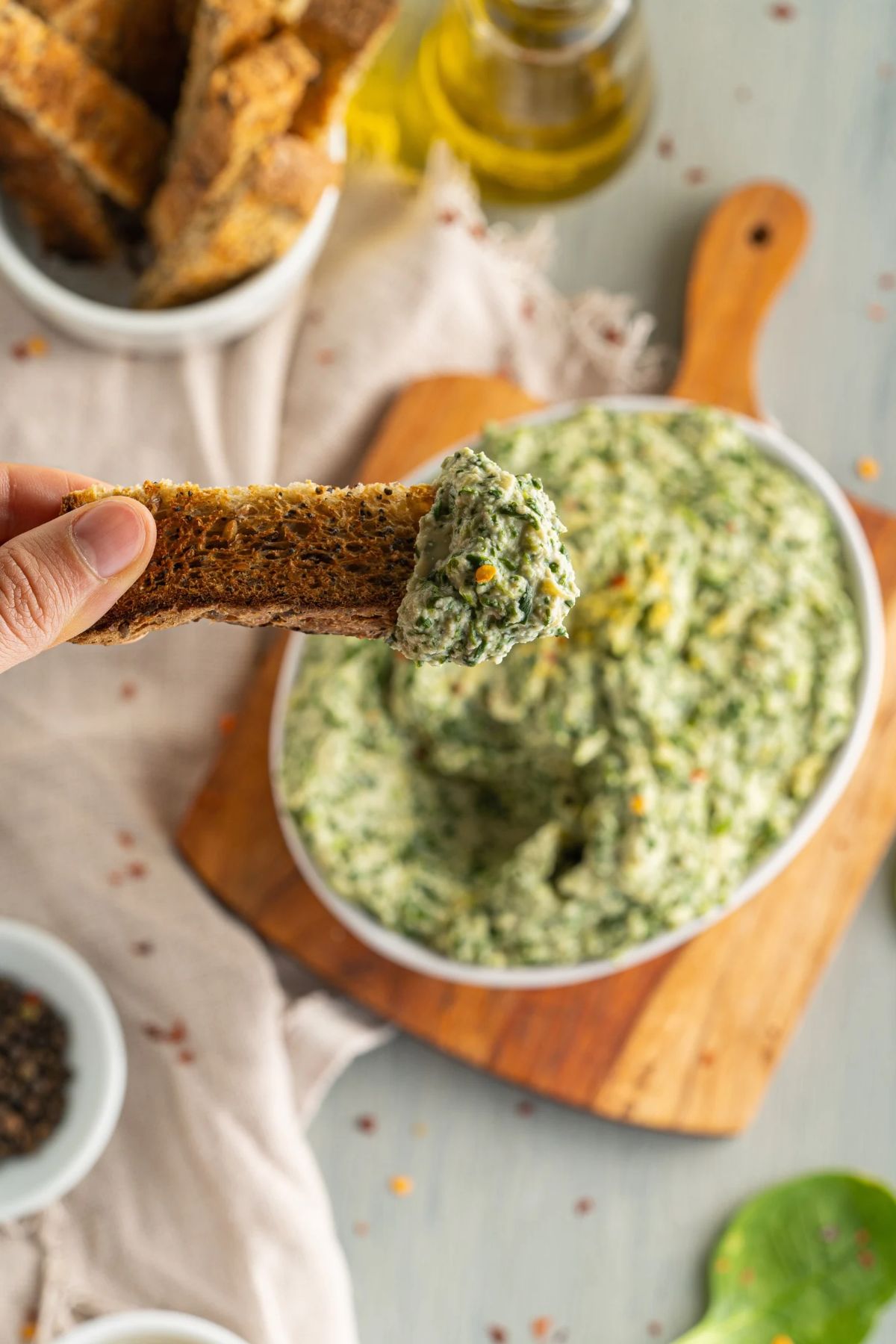 A hand holds a piece of toasted bread dipped into a serving of creamy spinach artichoke dip.